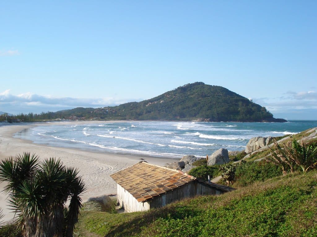 Praia da Ferrugem: uma praia, entornada por uma sutil região montanhosa esverdeada. Em primeiro plano, uma casa e um declive com grama verde.