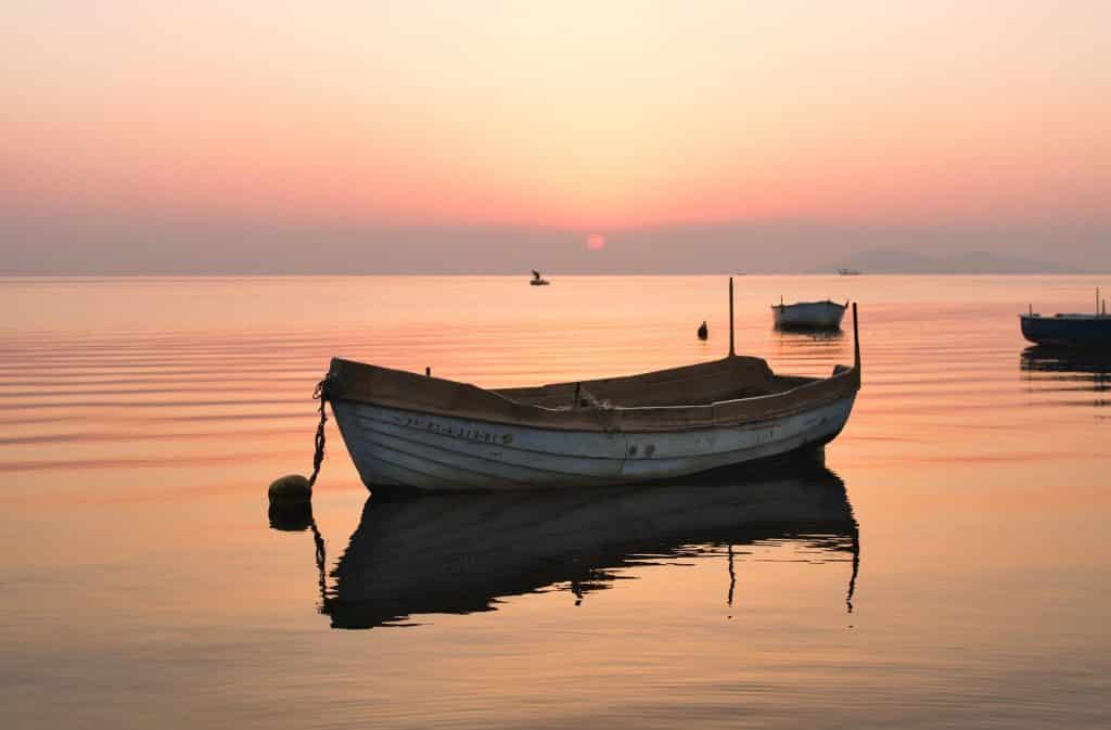 Um barco pequeno de coloração branco no meio de um rio. Ao fundo, outros barcos. Há também um pôr-do-sol.