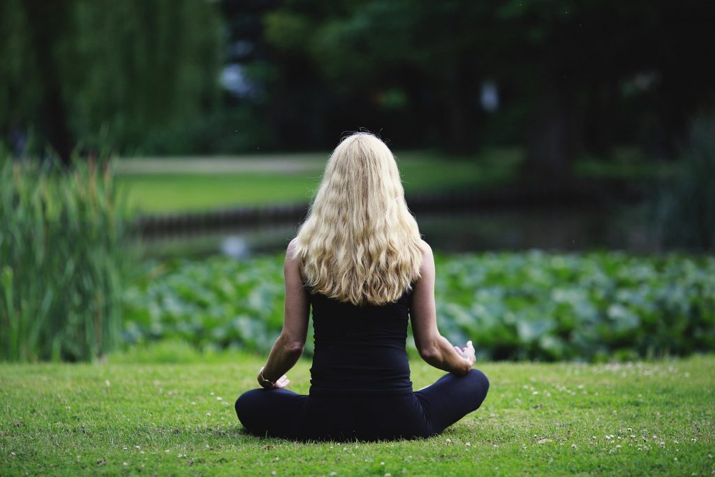 Uma mulher meditando num parque repleto de zonas verdes. Ao fundo, desfocado, um lago.