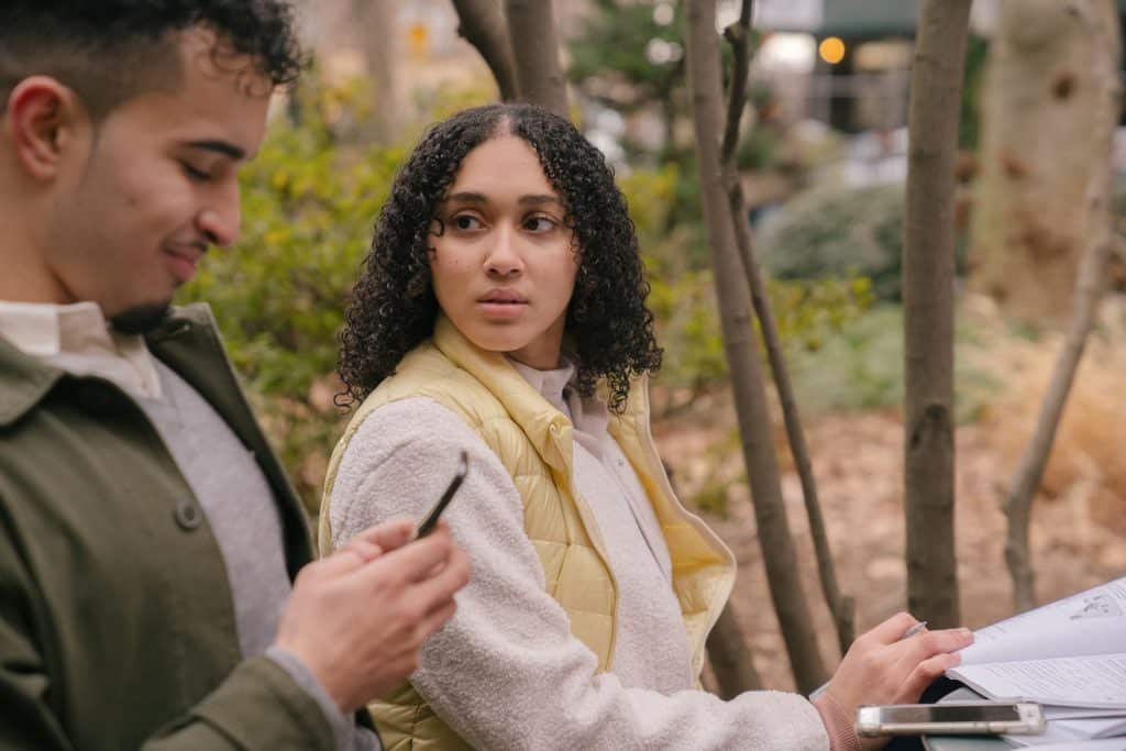 Um homem sorridente mexendo num celular e uma mulher olhando pra ele, aflitiva.