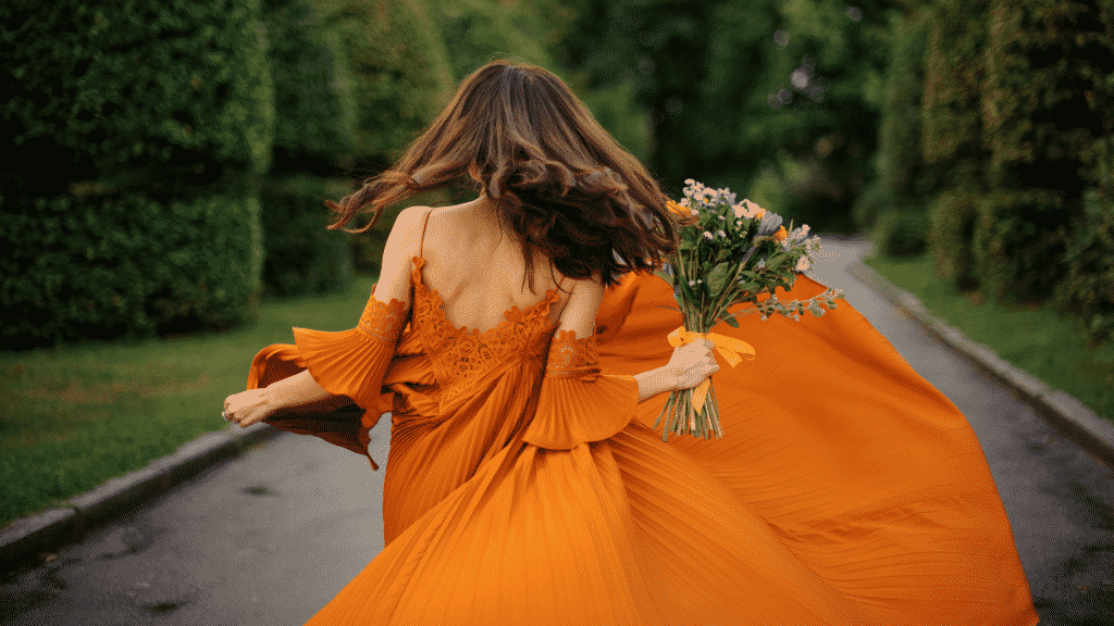 Mulher usando um longo vestido laranja e segurando um buquê de flores, de costas para a foto