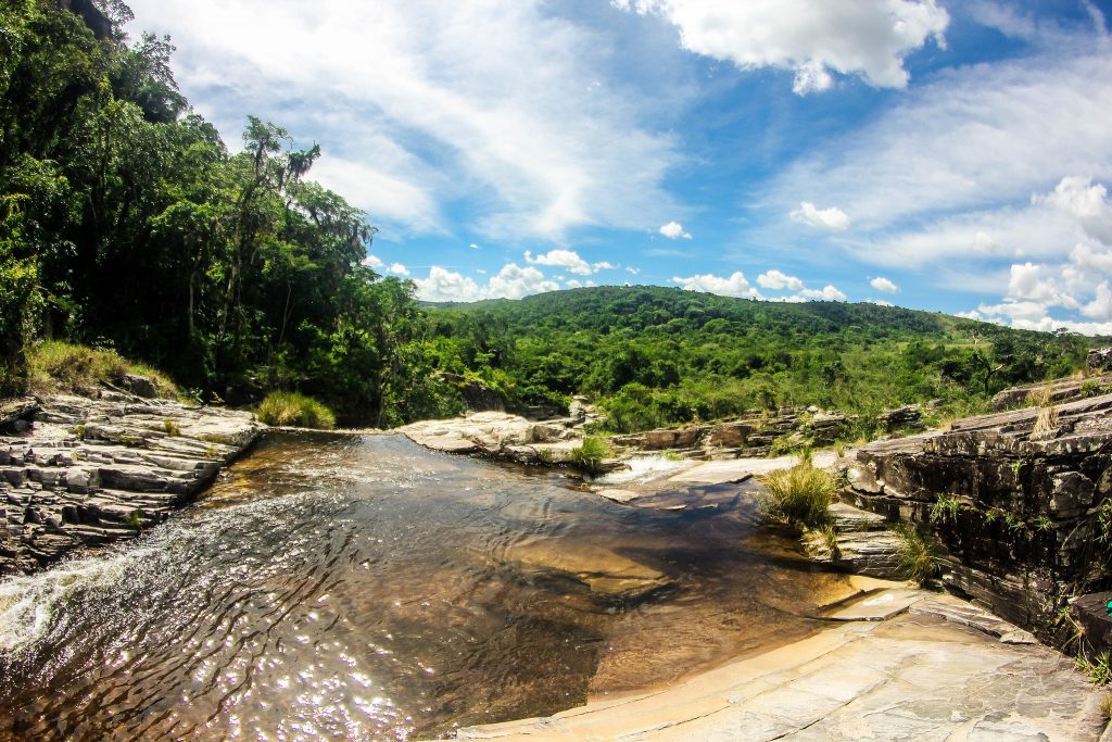 Uma pequena margem de uma cachoeira. Ao fundo, um céu azul repleto de nuvens e uma zona verde em árvores imensa.