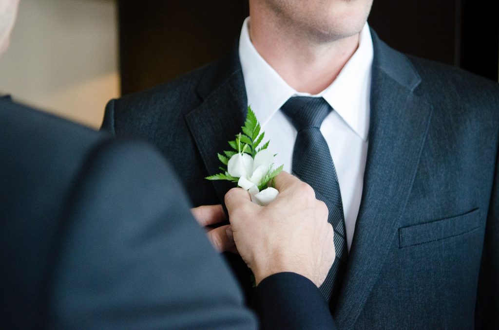 Ao fundo, um homem de terno. Em primeiro plano, embora situado com desfoque, vê-se um homem colocando um ramo de pequenas plantas sobre o terno do outro homem.