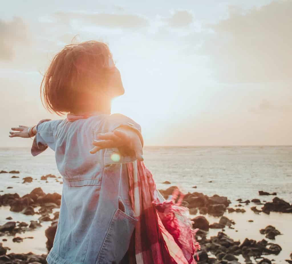 Uma mulher de braços erguidos contemplando um pôr-do-sol no mar.