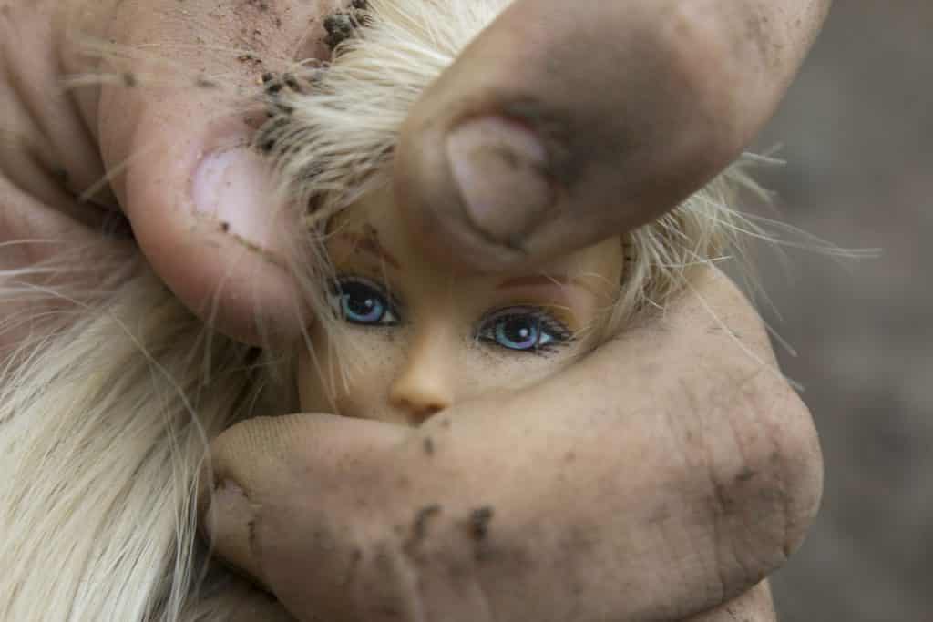 A cabeça de uma boneca feminina sendo apertada por uma mão possivelmente masculina.