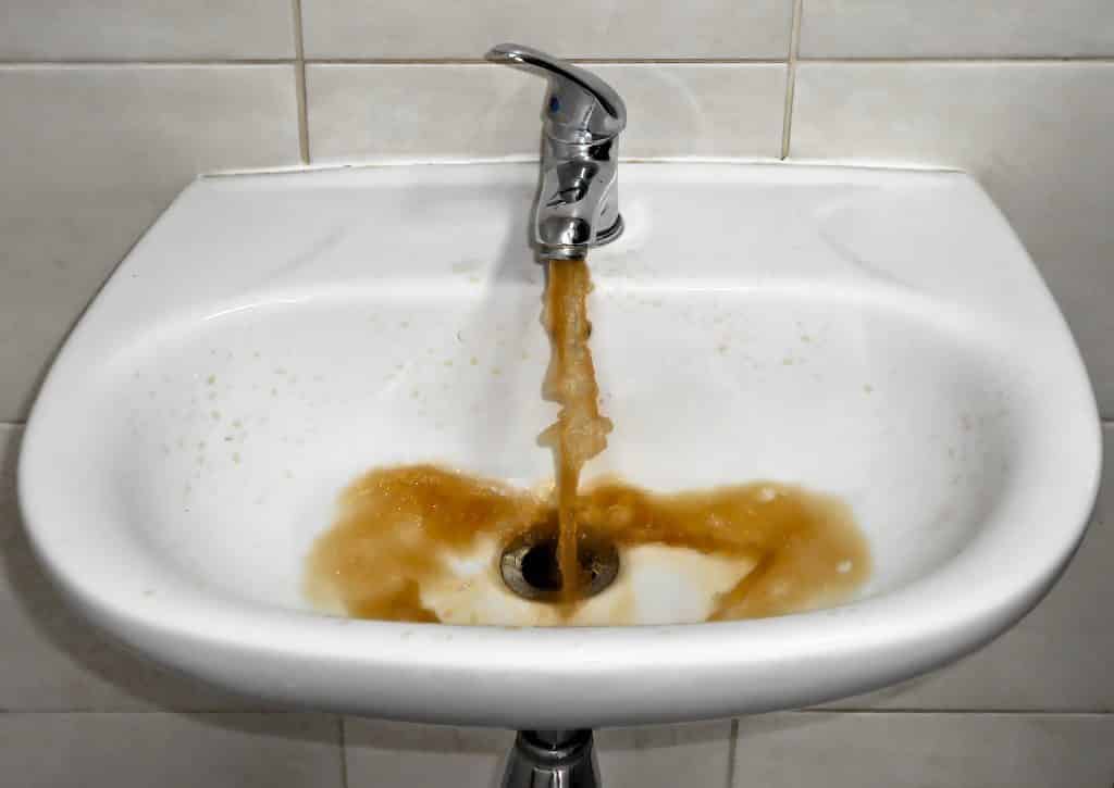 Uma torneira expelindo, numa pia, água suja de cor marrom.