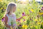 Uma garota branca de cabelos loiros em meio a um campo florido. Ela segura uma flor