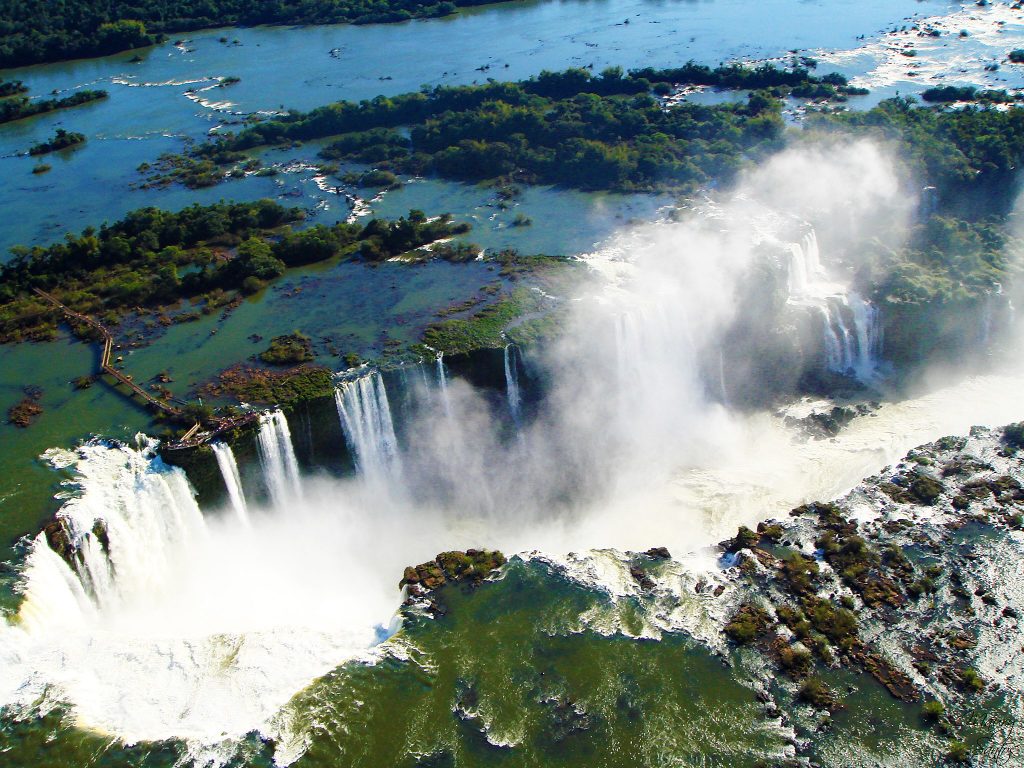 Uma visão aérea das passarelas aquáticas e das cataratas de Foz do Iguaçu. Grandes cachoeiras e, acima delas, zonas esverdeadas com águas rasas.