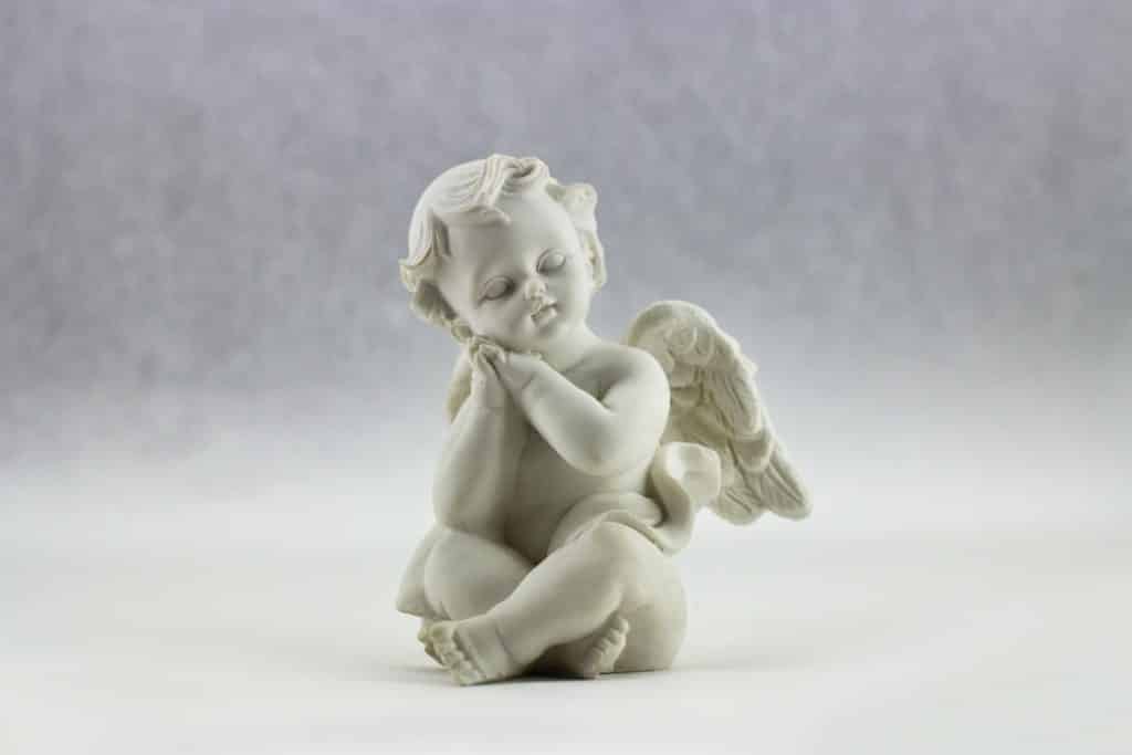 Uma estatua de um anjo com as mãos cruzadas. Ele debruça o rosto sobre elas e tem asas nas costas.