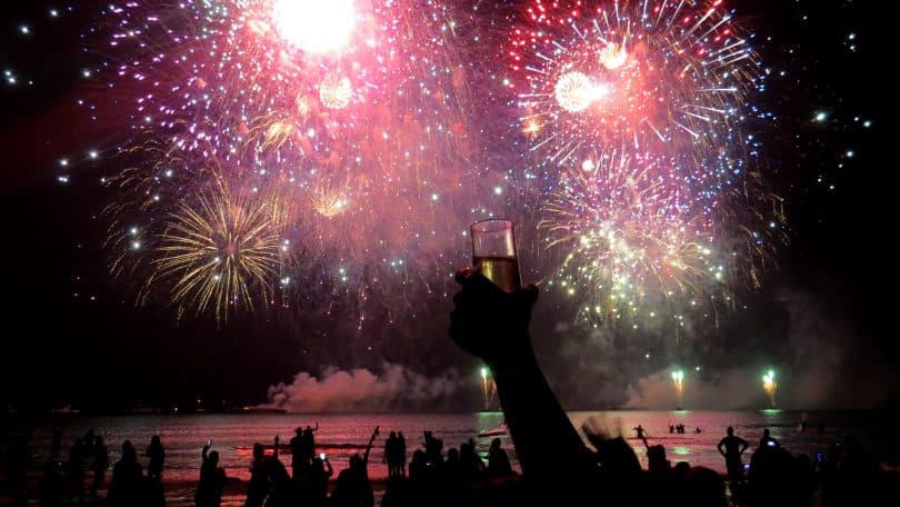 Fogos de artifício luminosos em meio à escuridão. Embaixo deles e em primeiro plano, pessoas de braços erguidos e comemorando.