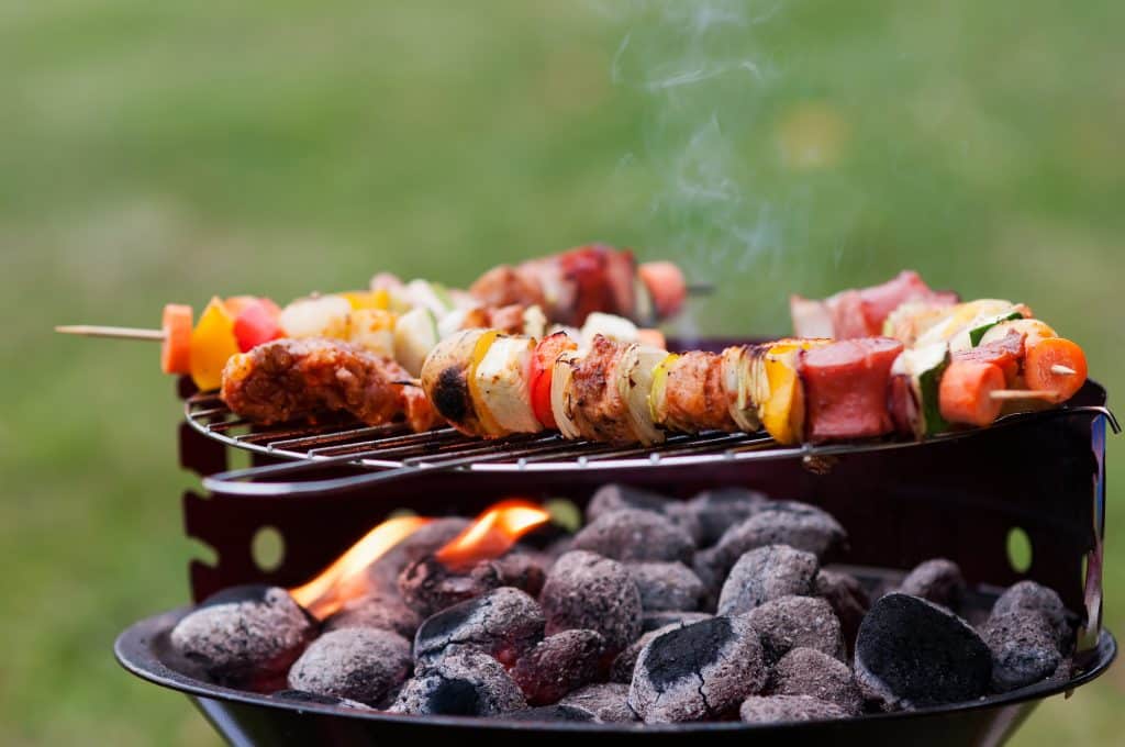 Espetos de carnes e alimentos variados sendo feitos em uma churrasqueira. Embaixo dos espetos, subjazendo a uma grade, carvão.