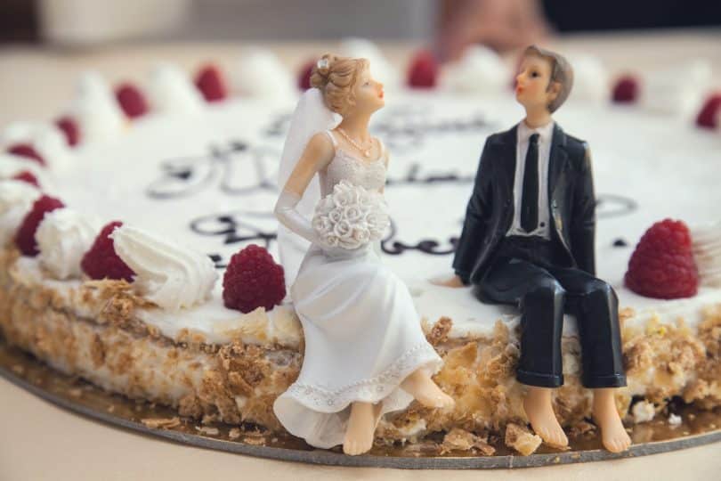 Sobre um bolo, miniaturas de noivos, sendo uma representante de um homem e a outra de uma mulher.