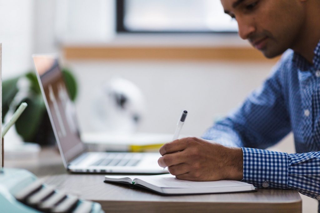 Um homem escrevendo sobre um caderno posto numa mesa. Nessa mesa, desfocado, um notebook.