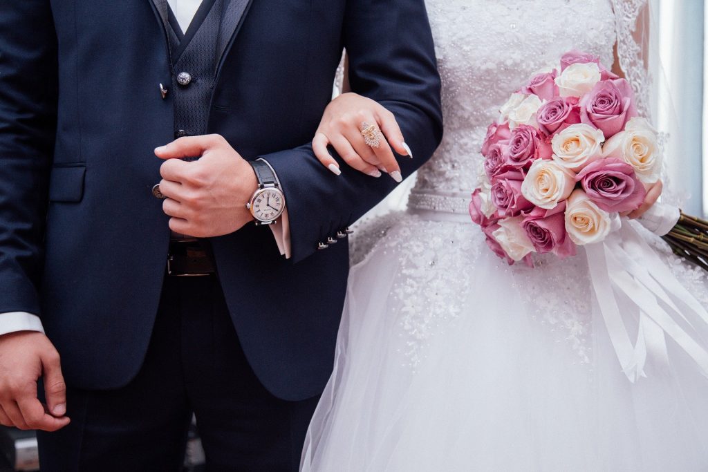 Um homem trajado de terna e, à sua esquerda, uma mulher vestindo um vestido e segurando um buquê de flores. A mão esquerda da mulher está sobre o braço esquerdo do homem.