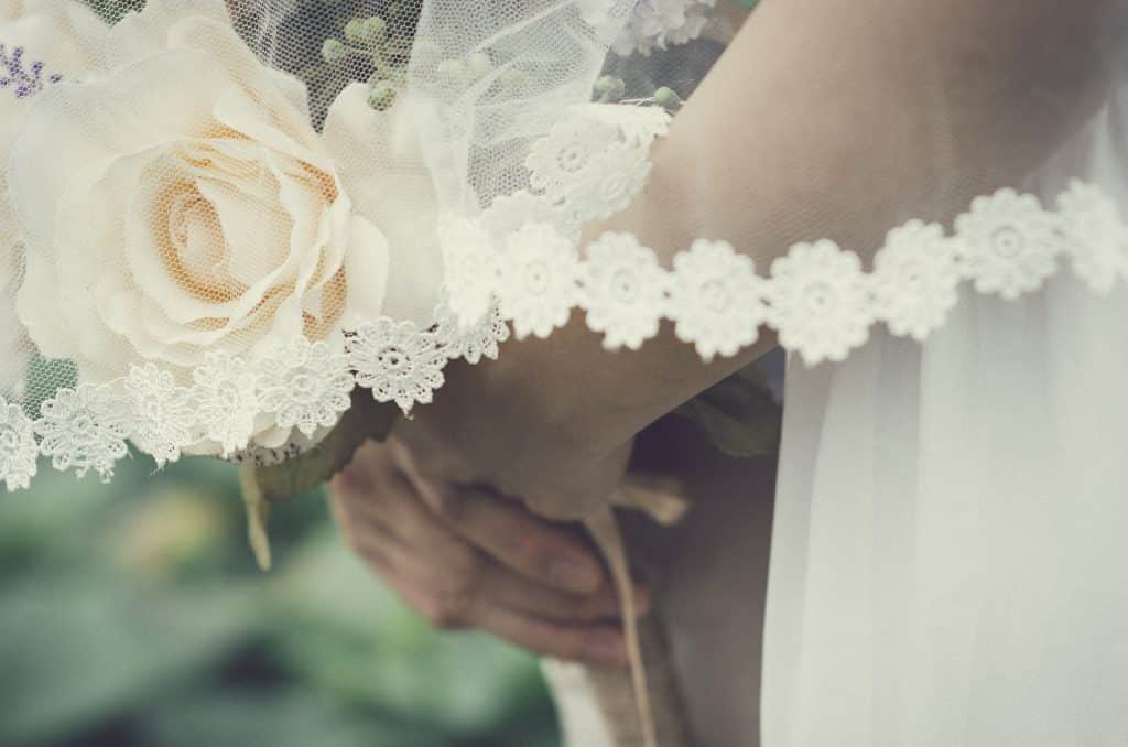 Braços e mãos supostamente femininas segurando um buquê de flores. Sobre os braços, um véu branco caindo sutilmente.