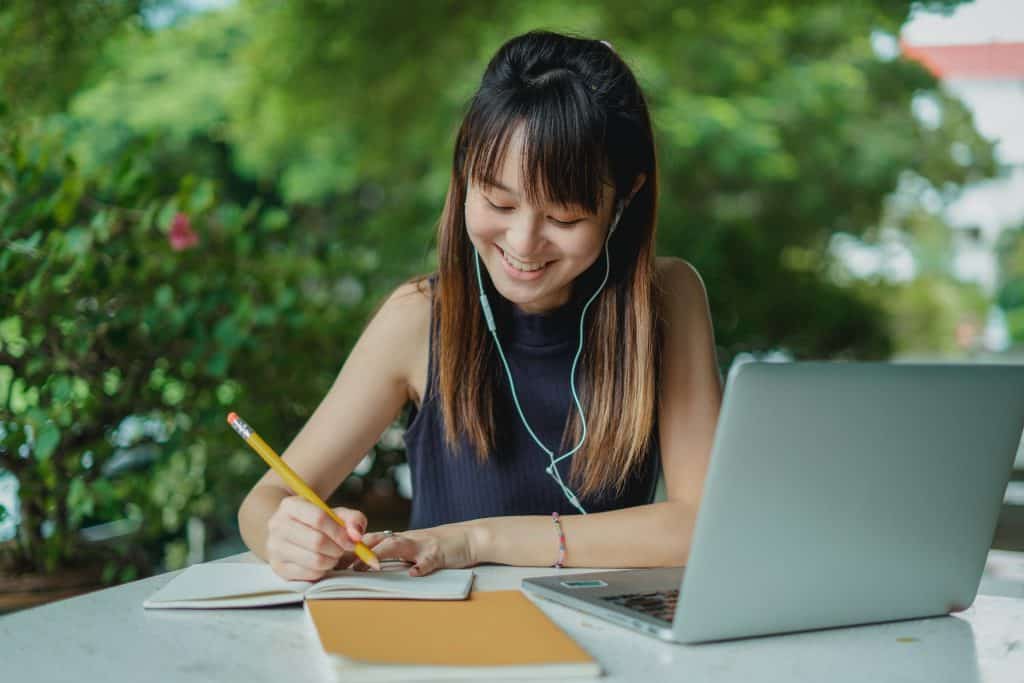 Uma mulher asiática fazendo anotações em um caderno. Em sua mesa, outro caderno e um notebook. Ela sorri e usa fones de ouvido.