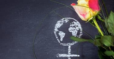 Uma ilustração em giz, numa lousa, de um globo terrestre mesclado com o símbolo que representa o feminino. Ao lado, uma flor.