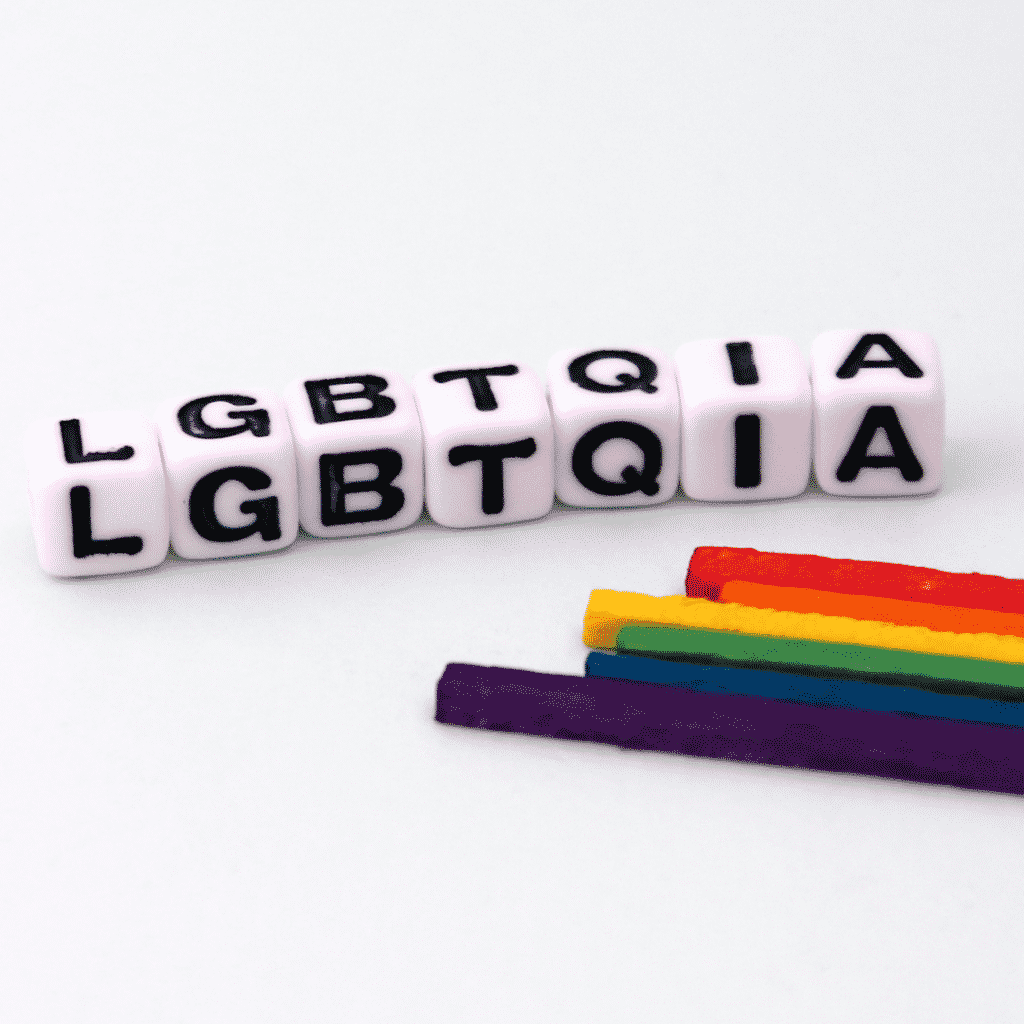 7 dados com as letras L G B T Q I A. À direita, pedaços retangulares representando as cores LGBTQIA+, roxo, azul, verde, amarelo, laranja e vermelho.