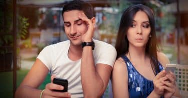 Um homem mexendo num celular e à esquerda uma mulher bisbilhotando o celular dele.