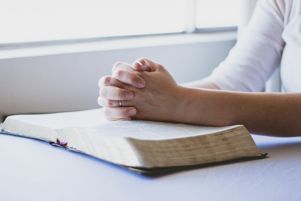 Uma pessoa com os dedos entrelaçados em um gesto de oração. Ela debruça suas mãos sobre uma provável bíblia.