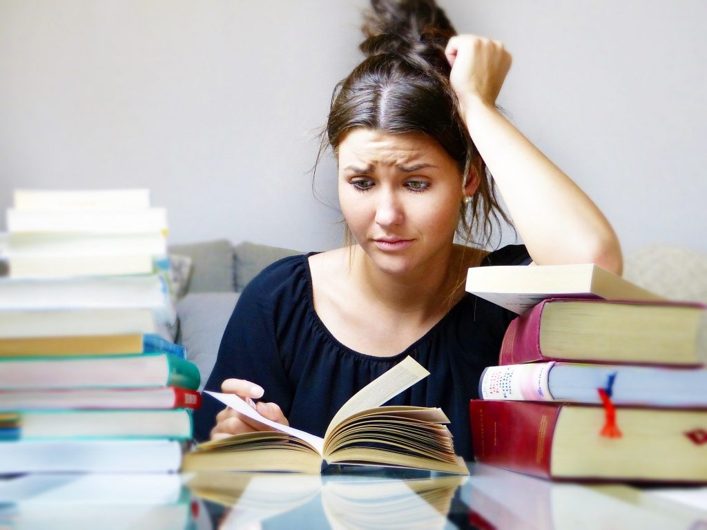 Uma mulher com aparente descontentamento lendo um livro. À sua direita e à sua esquerda, duas pilhas de livros.