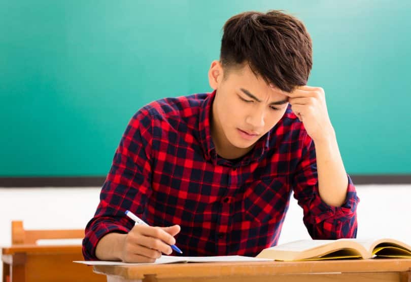 Um homem escrevendo em um caderno numa mesa. Ele parece estressado.