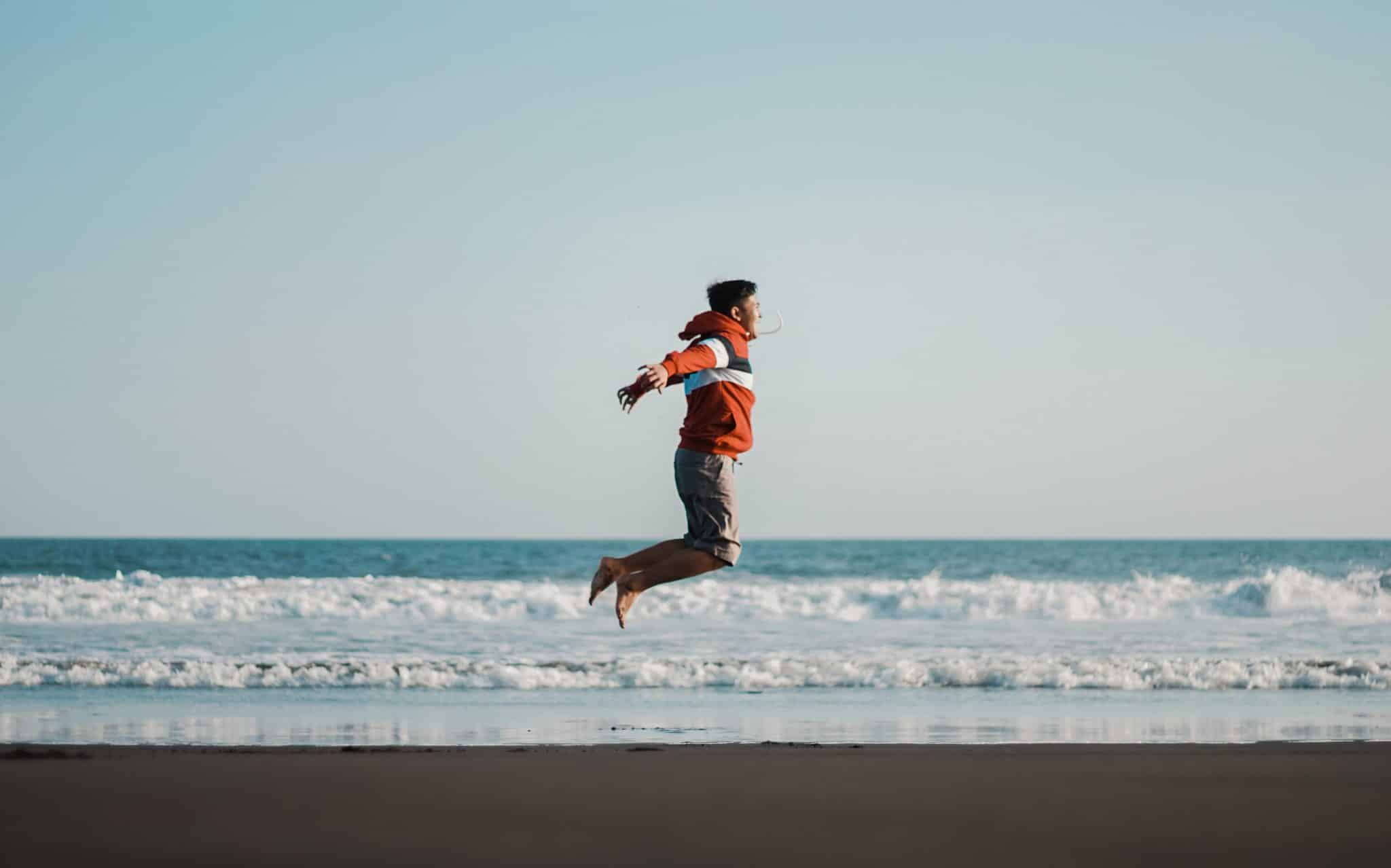 Um homem pulando em uma praia. Em plano posterior, o mar.