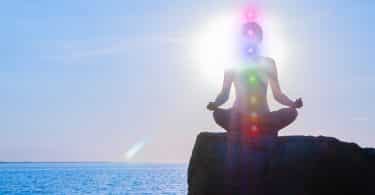 Mulher está meditando com brilhantes sete chakras na pedra ao pôr do sol.
