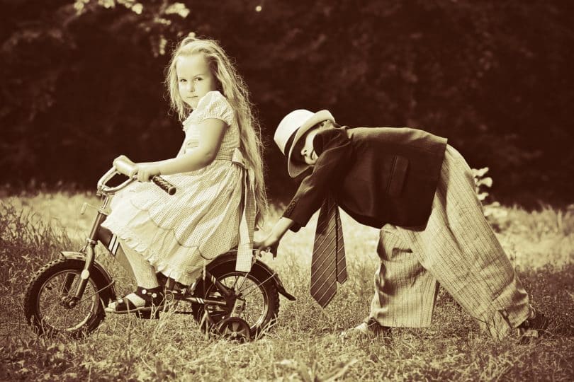 Menina e menino brancos brincando de bicicleta num campo.