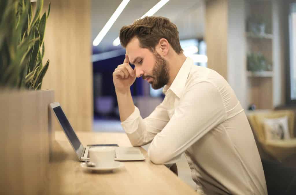 Um homem de roupa social debruçando seus braços sobre uma mesa com um notebook. Ele parece consternado.