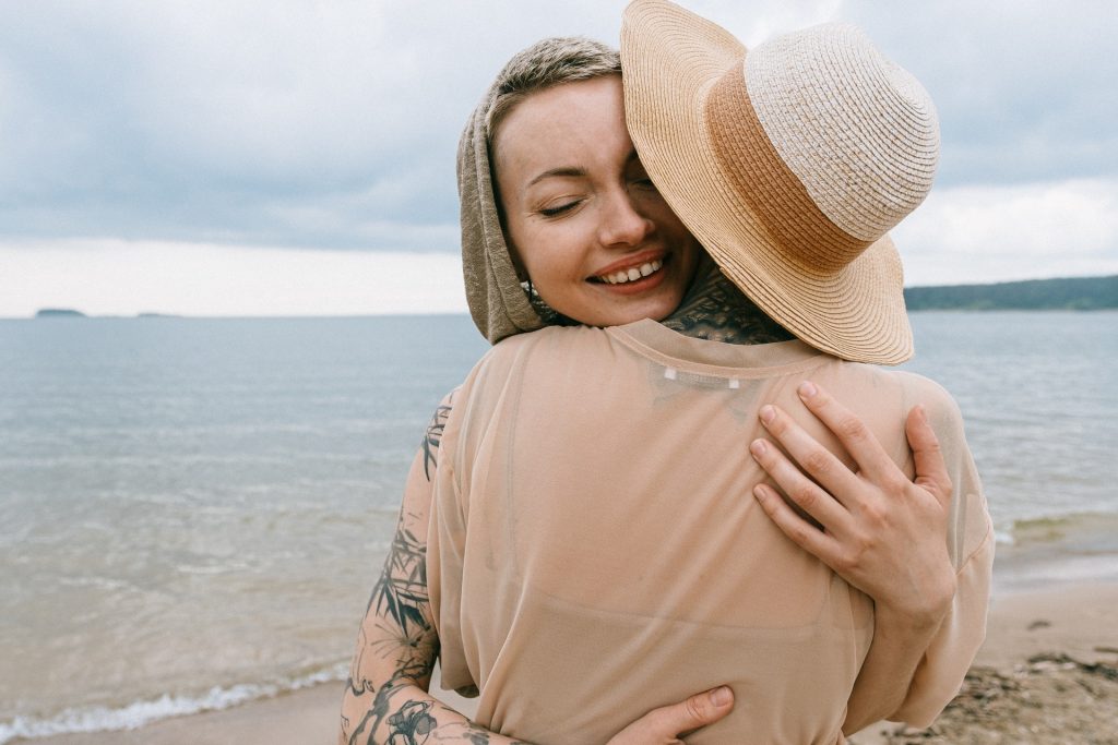 Uma mulher abraçando uma pessoa em frente à uma praia.