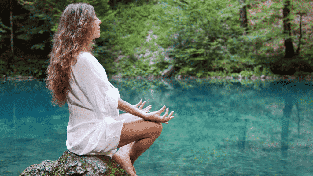 Mulher sentada em uma pedra na beira de um rio, meditando e de olhos fechados