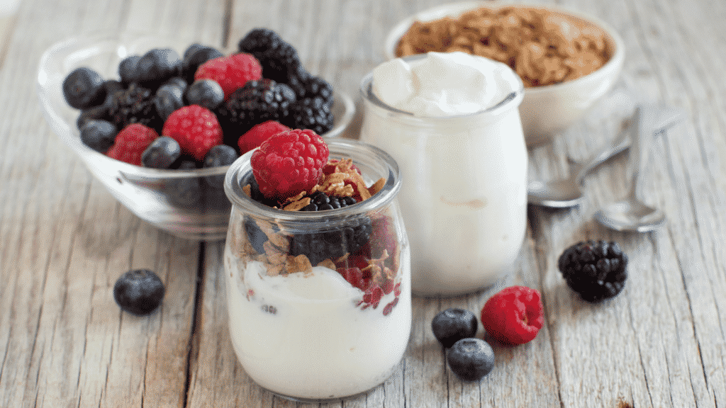 Iogurte branco, granola e frutas em recipientes de vidro, em cima de uma mesa de madeira