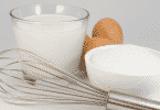 Leite em um copo de vidro, ovos, açúcar e um batedor manual de ovos sobre uma mesa