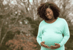 Mulher gestante sorrindo e segurando a barriga. Atrás, uma árvore