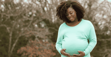 Mulher gestante sorrindo e segurando a barriga. Atrás, uma árvore