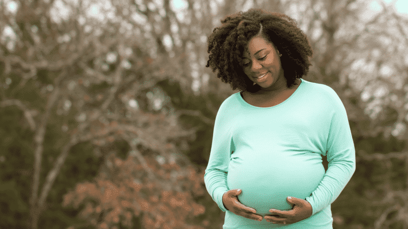 Mulher gestante sorrindo e segurando a barriga. Atrás, uma árvore