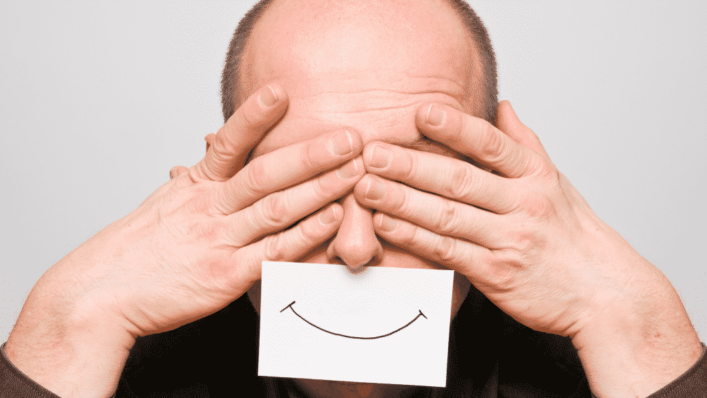 Homem tapando os olhos com as mãos. Um papel com um sorriso desenhado está colado em cima da sua boca