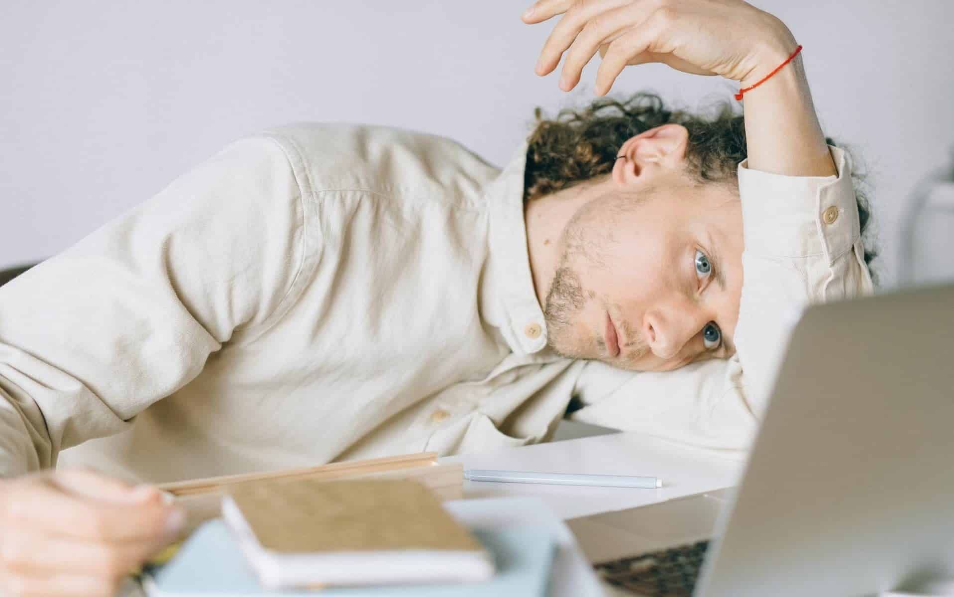 Um homem debruçando sua cabeça sobre o seu braço esquerdo. Ele olha para um notebook e tem um semblante de estresse.