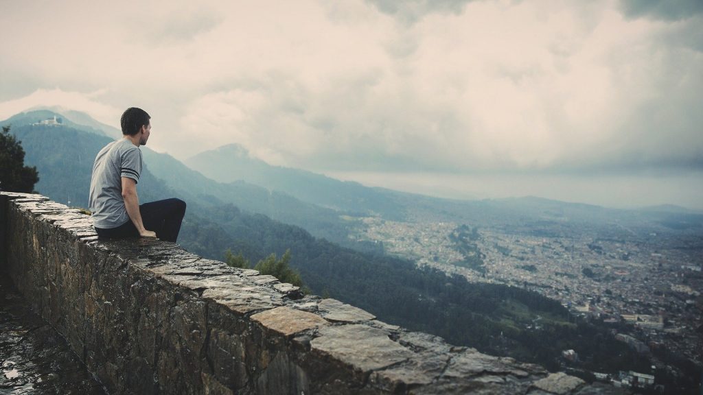 Um homem sentado em um muro contemplando, do alto, uma vista panorâmica e horizontal de uma zona verde e altamente montanhosa.