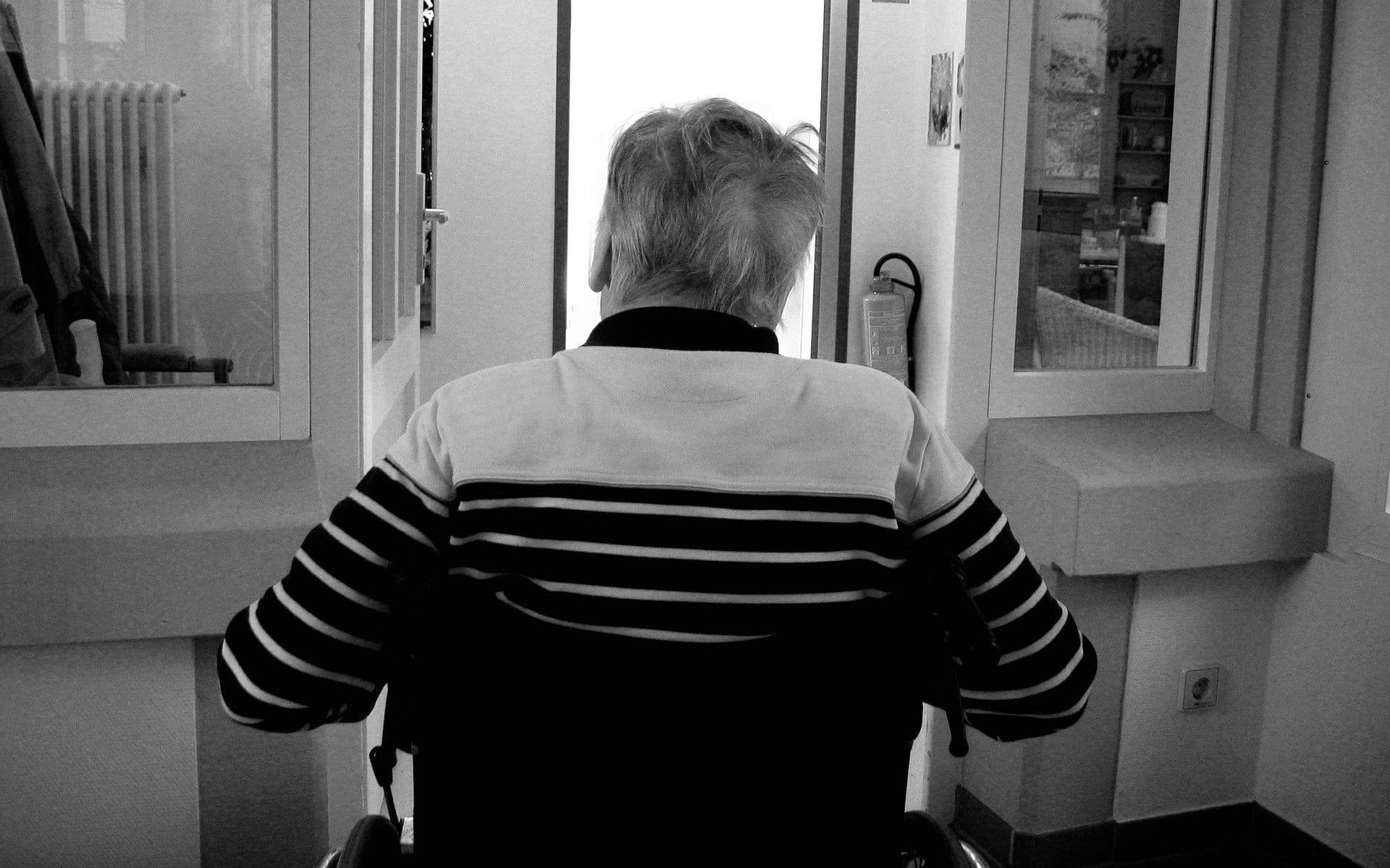 Uma foto em preto e branco: um idoso visto de costas sentado numa cadeira de rodas.