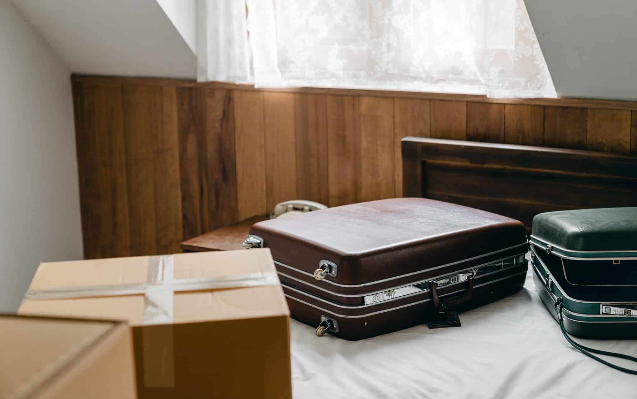 Sobre uma cama, malas e caixas de papelão seladas. No fundo, uma janela emitindo luz através de uma fresta.