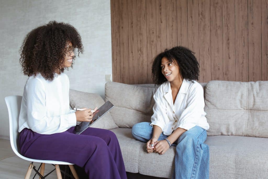 Duas mulheres negras conversando. À esquerda, uma sentada numa cadeira, à direita, outra sentada num sofá. Elas sorriem.