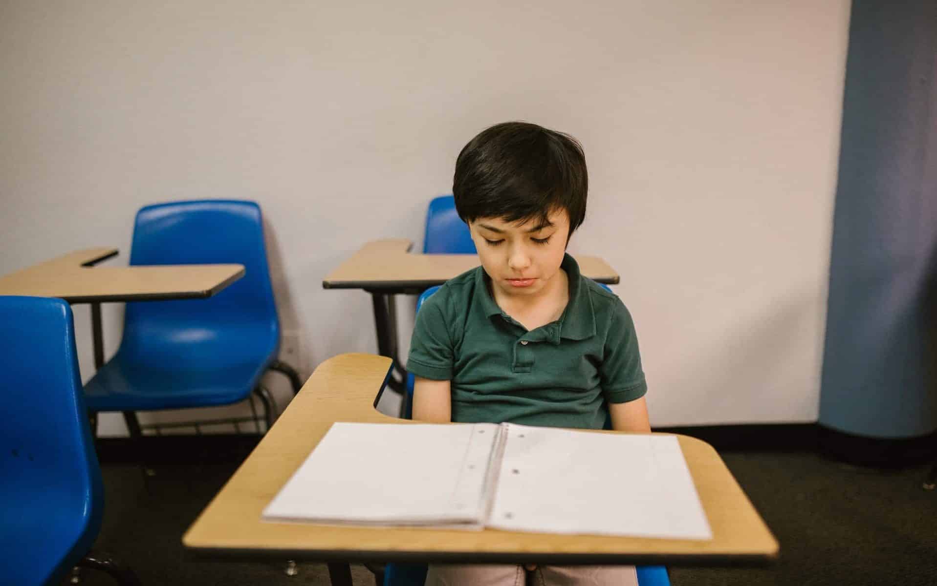 Um garoto sentado próximo à uma mesa de escola. Ele olha para um caderno em branco posto sobre a mesa e parece frustrado.
