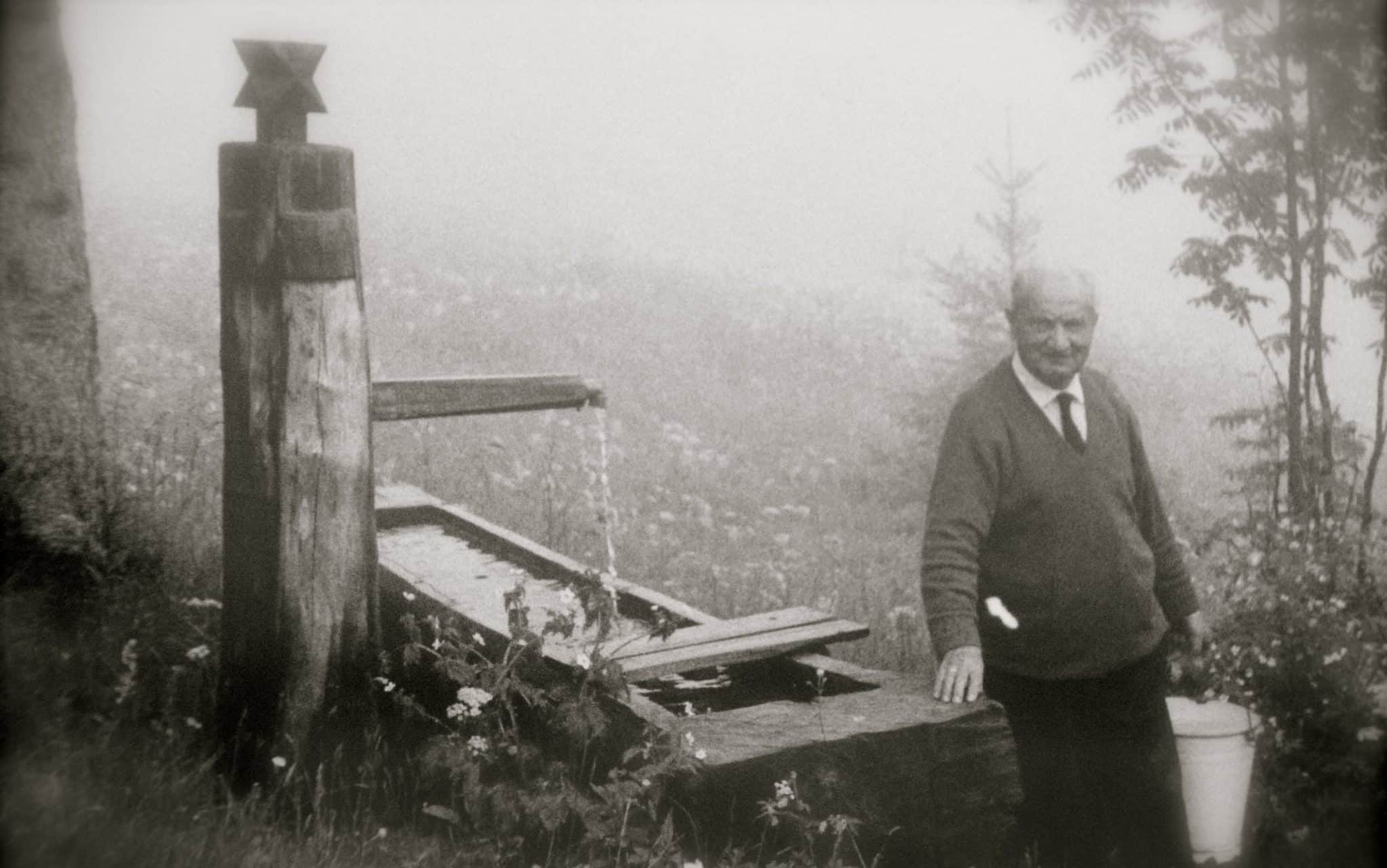 Uma fotografia de Heidegger. Ele está situado próximo a um poço, carregando um balde d'água e sorrindo.