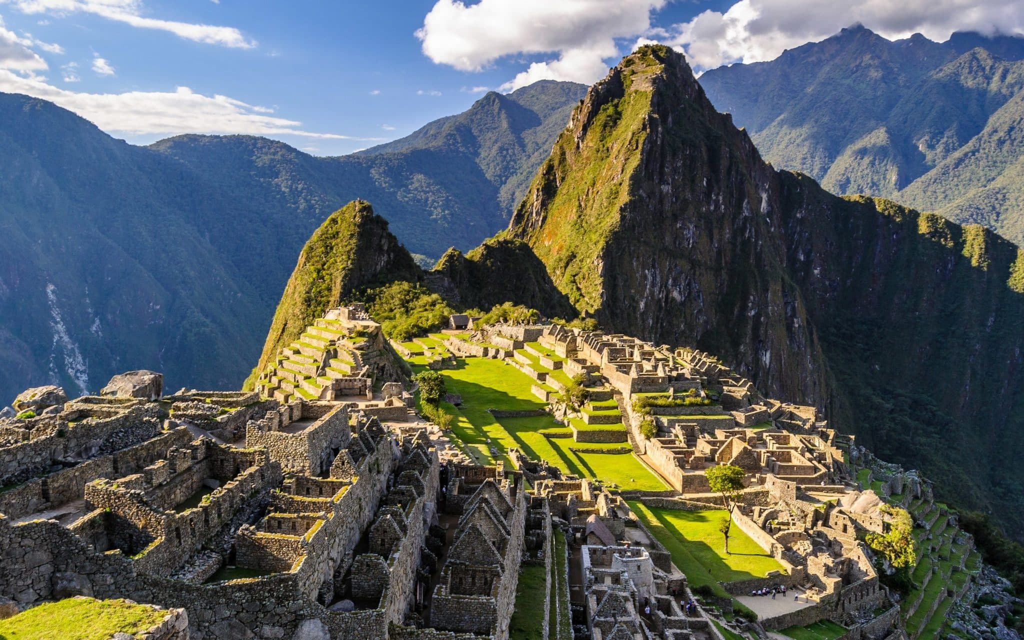 Picos montanhosos e cumes verdes. Próximo a eles, construções que assemelham-se a muros. Ao fundo, mais grandes picos.