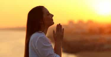 Uma mulher ergue as mãos fazendo um gesto de oração. Ao fundo, um sol poente.