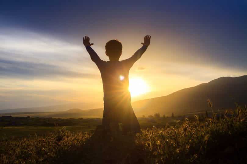 Um garoto erguendo as mãos em frente ao por do sol