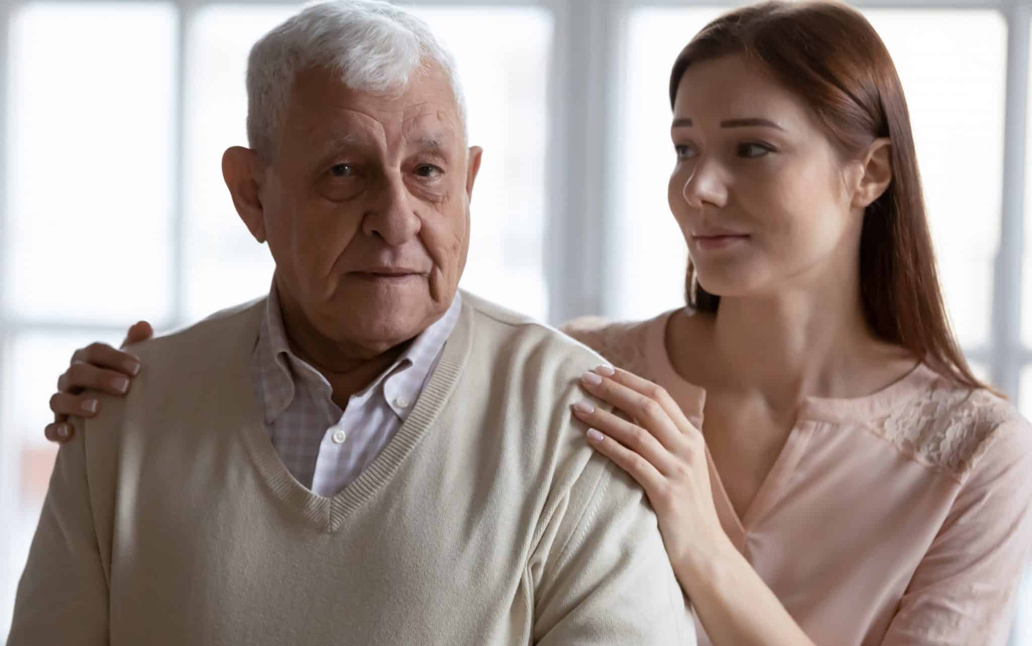 Um homem idoso e uma mulher ruiva colocando as mãos nos seus ombros.