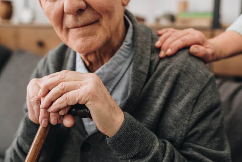 Um idoso segurando uma bengala. Parte do seu rosto sendo exibido e, no seu ombro, uma mão.